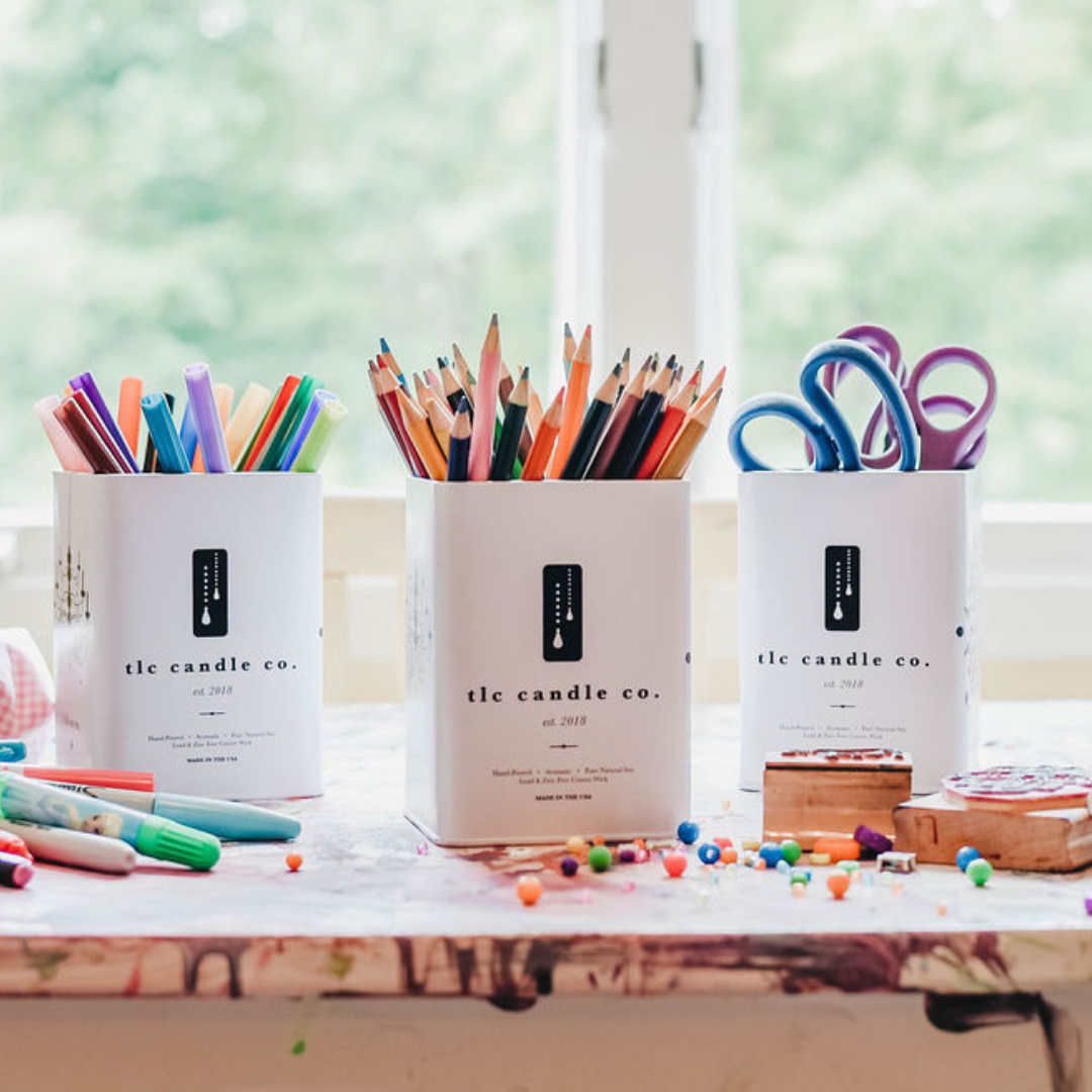 Three containers from 'tle candle co.' filled with stationery items on a desk.