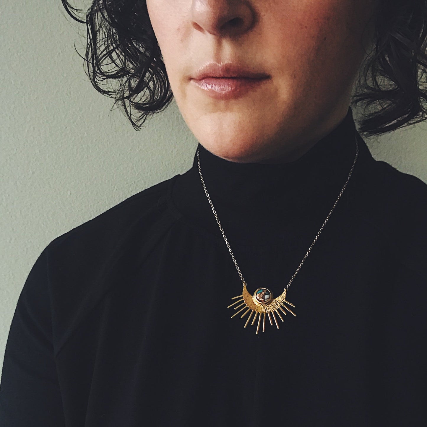 Person wearing a black turtleneck and a gold necklace with a sun design against a neutral background