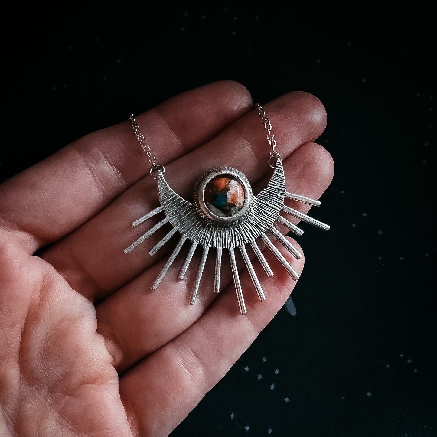 Silver necklace with a sunburst design held in a hand against a dark background