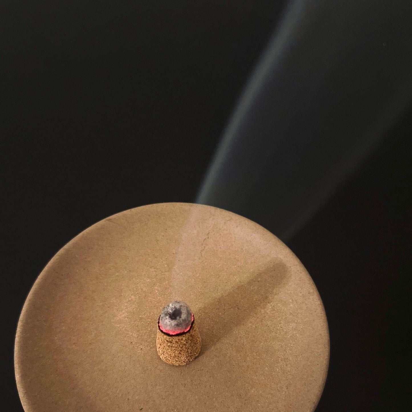 Incense cone burning on a round brown base with smoke rising against a black background