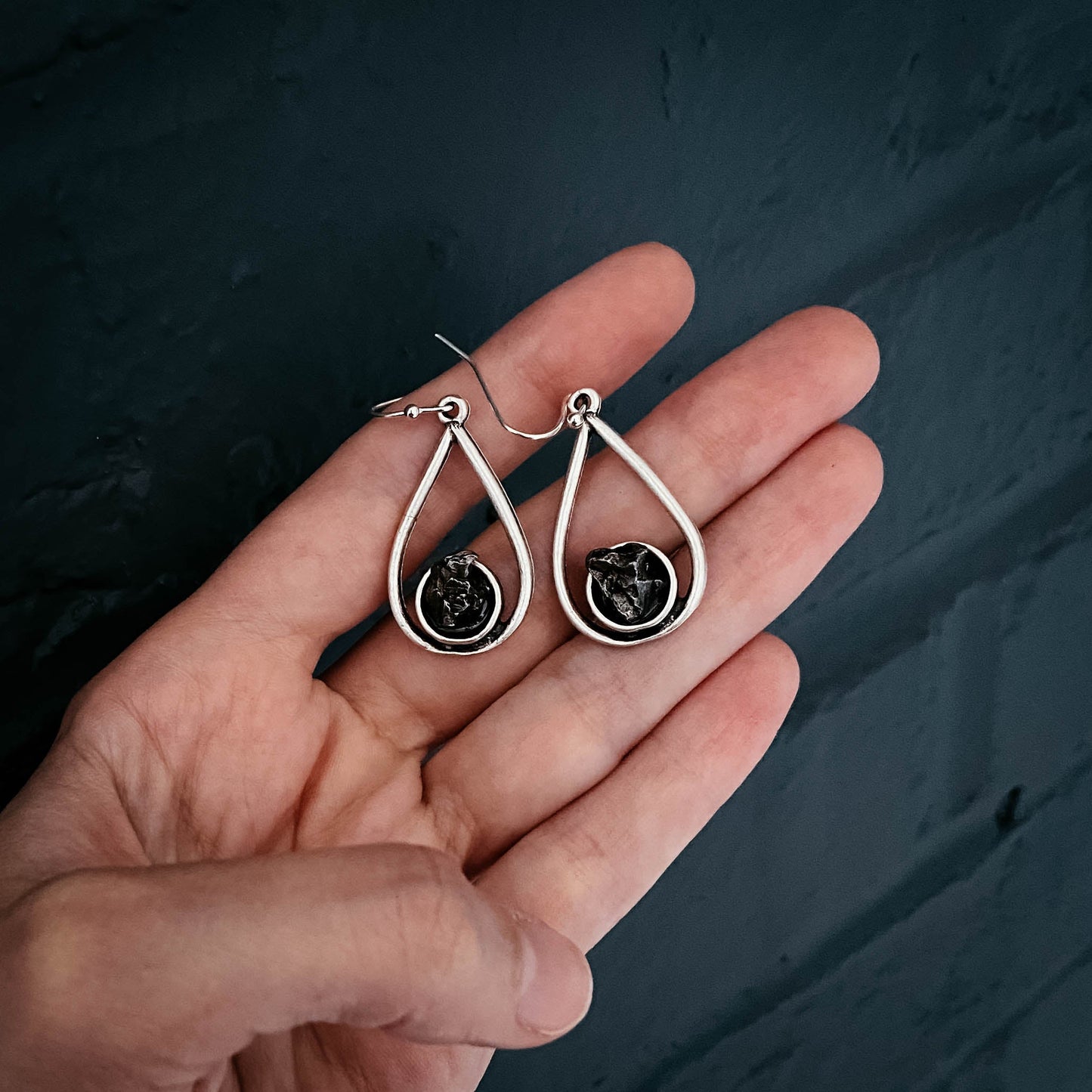 Silver teardrop earrings with black stones held in a hand against a dark background