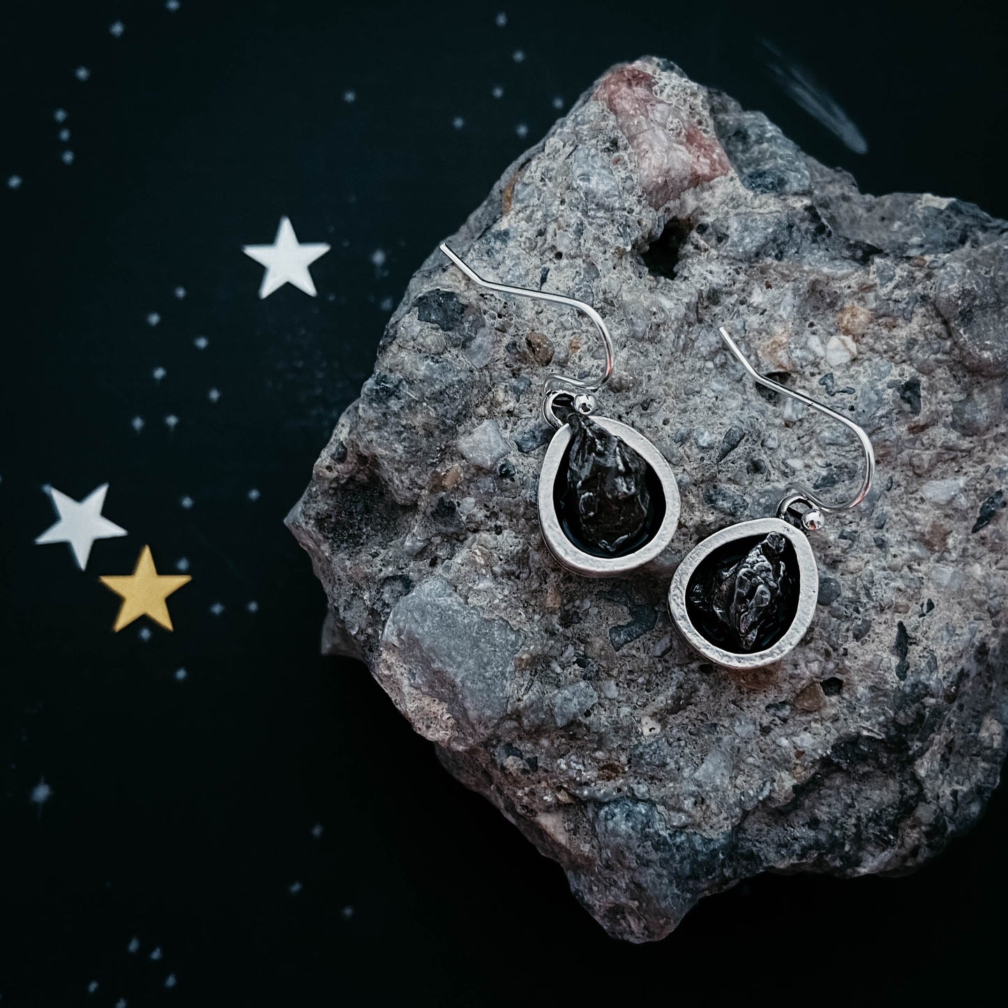 Silver earrings with black stones on a textured rock surface with star decorations.