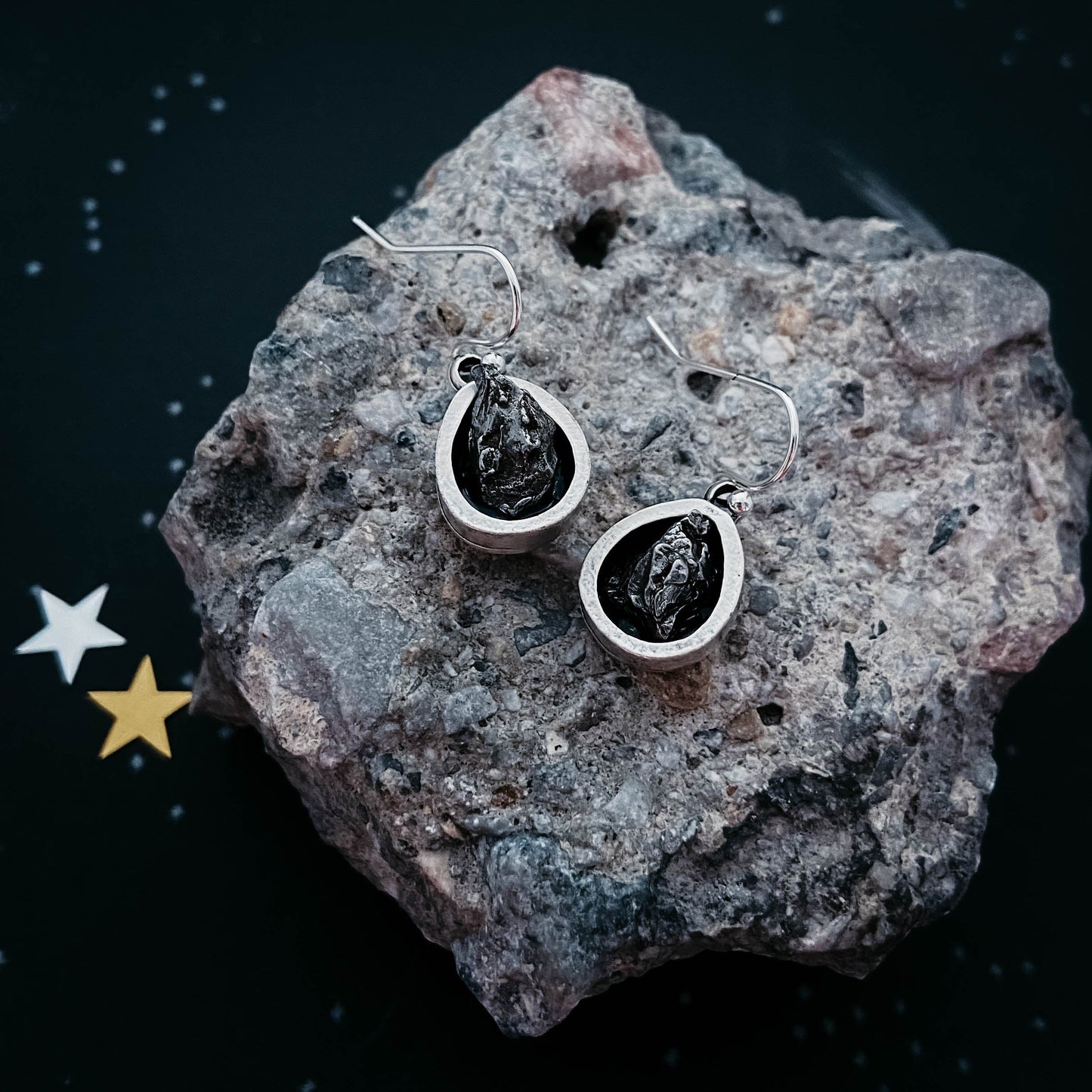 Silver earrings with black stones on a textured rock surface with star decorations.