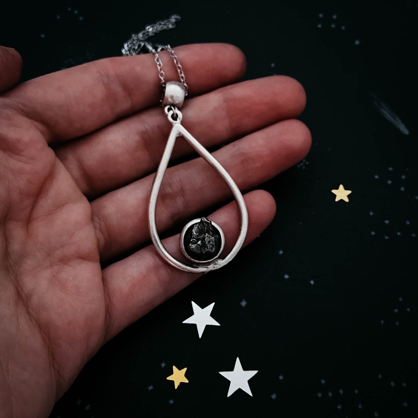 Silver teardrop-shaped pendant on a chain held in a hand with decorative stars on a dark background