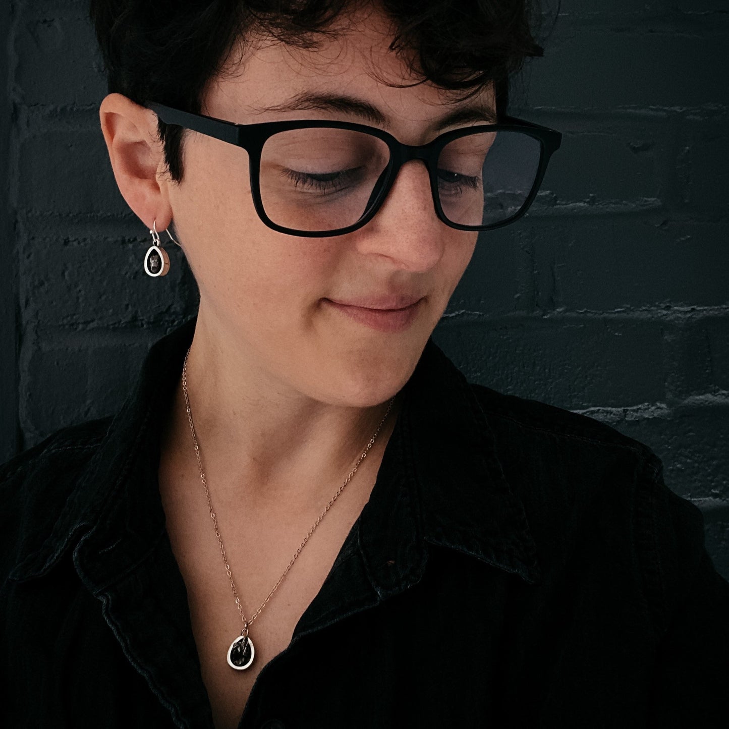 Person wearing black glasses and a black shirt against a dark brick wall