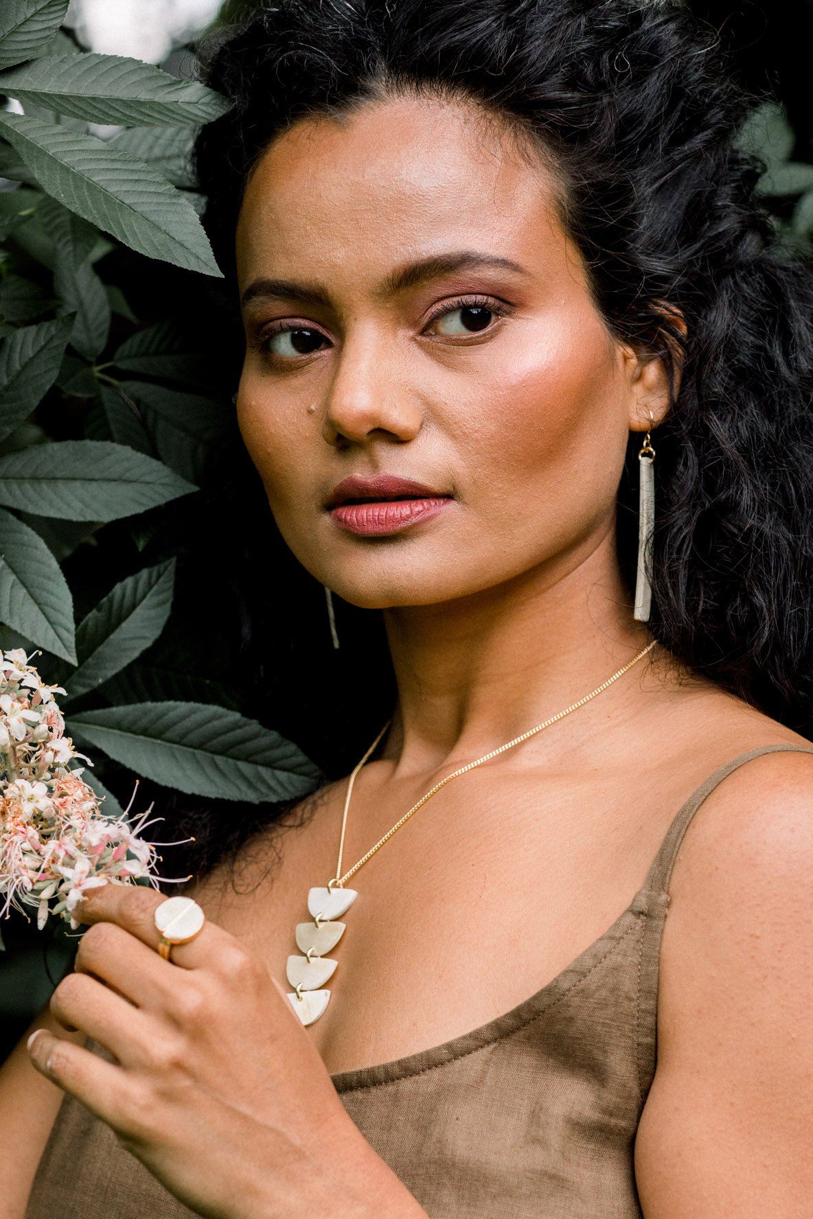 Woman with dark hair and earrings, wearing a necklace and ring, surrounded by greenery.