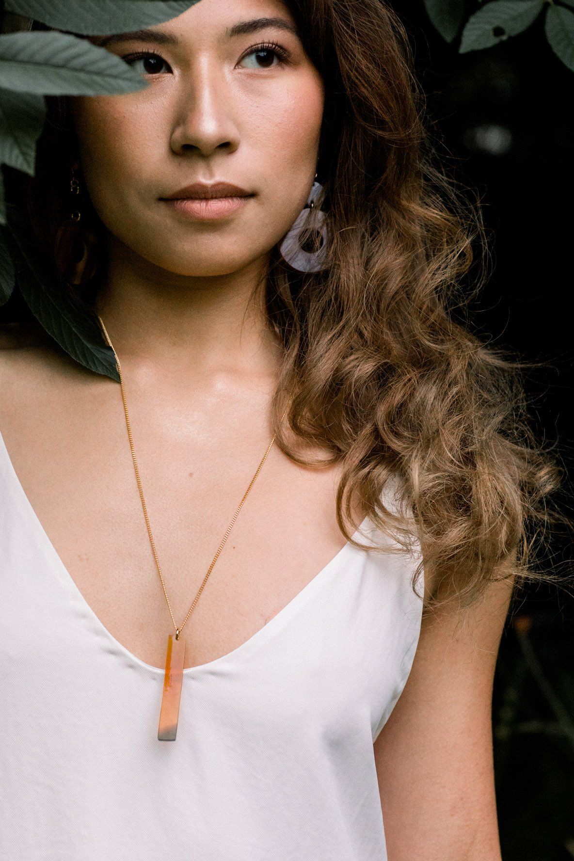 Woman wearing a white top and a necklace with a wooden pendant, surrounded by greenery.