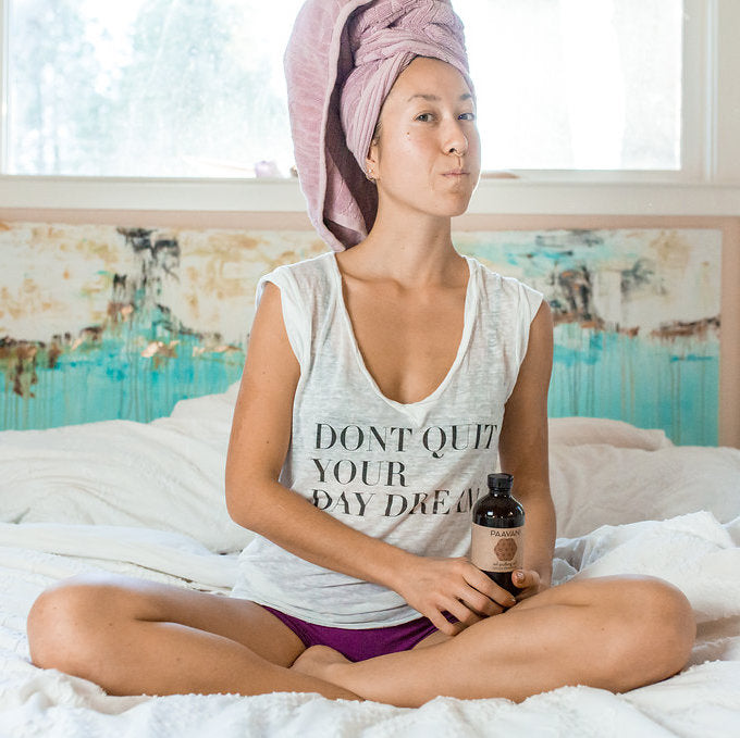 Woman sitting on a bed with a towel wrapped around her head, holding a bottle, in a room with a colorful abstract painting on the wall.