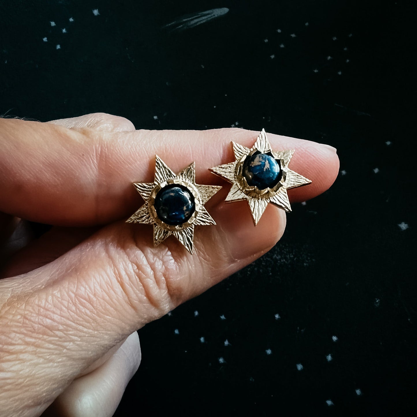 Gold star-shaped earrings with central stones held between fingers against a dark background