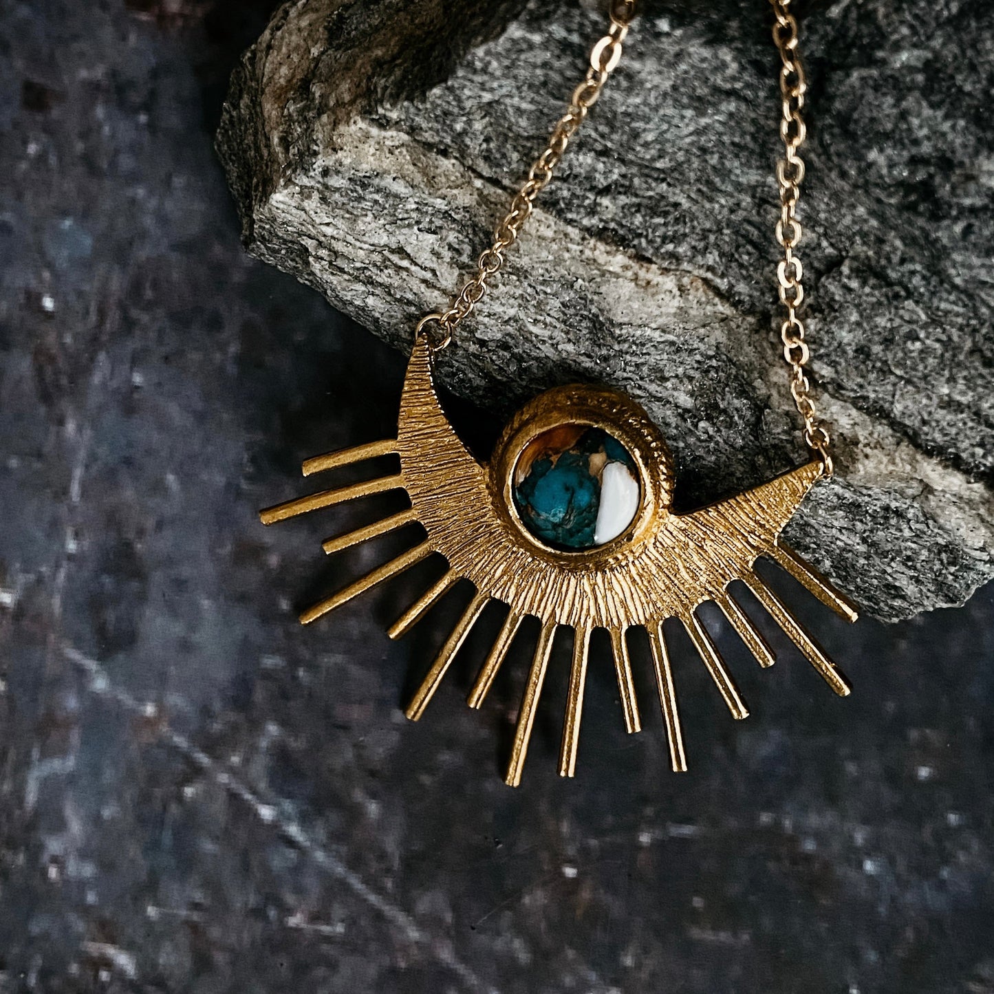 Gold necklace with a sun-shaped pendant featuring a blue gemstone on a dark stone background