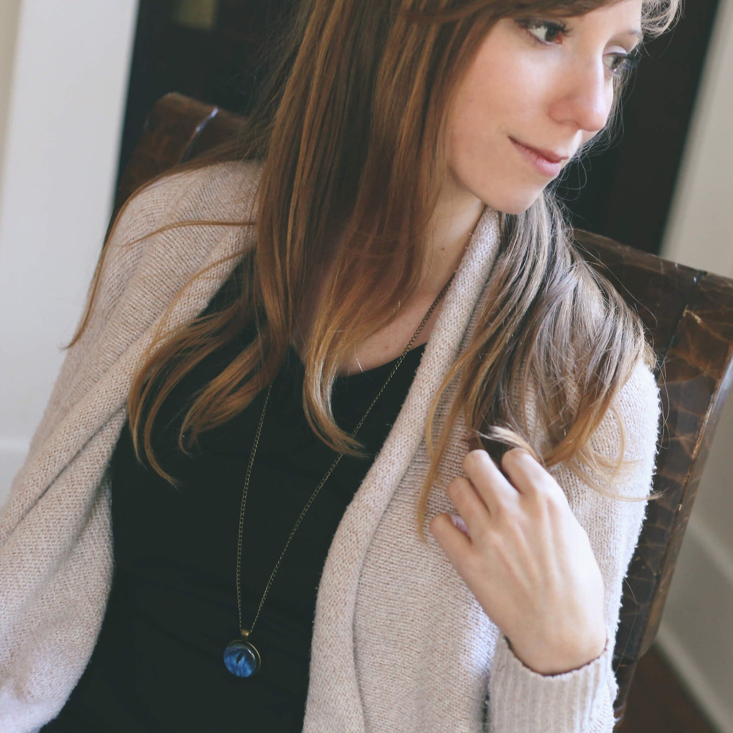 Woman wearing a beige cardigan and black top with a blue pendant necklace, sitting indoors.