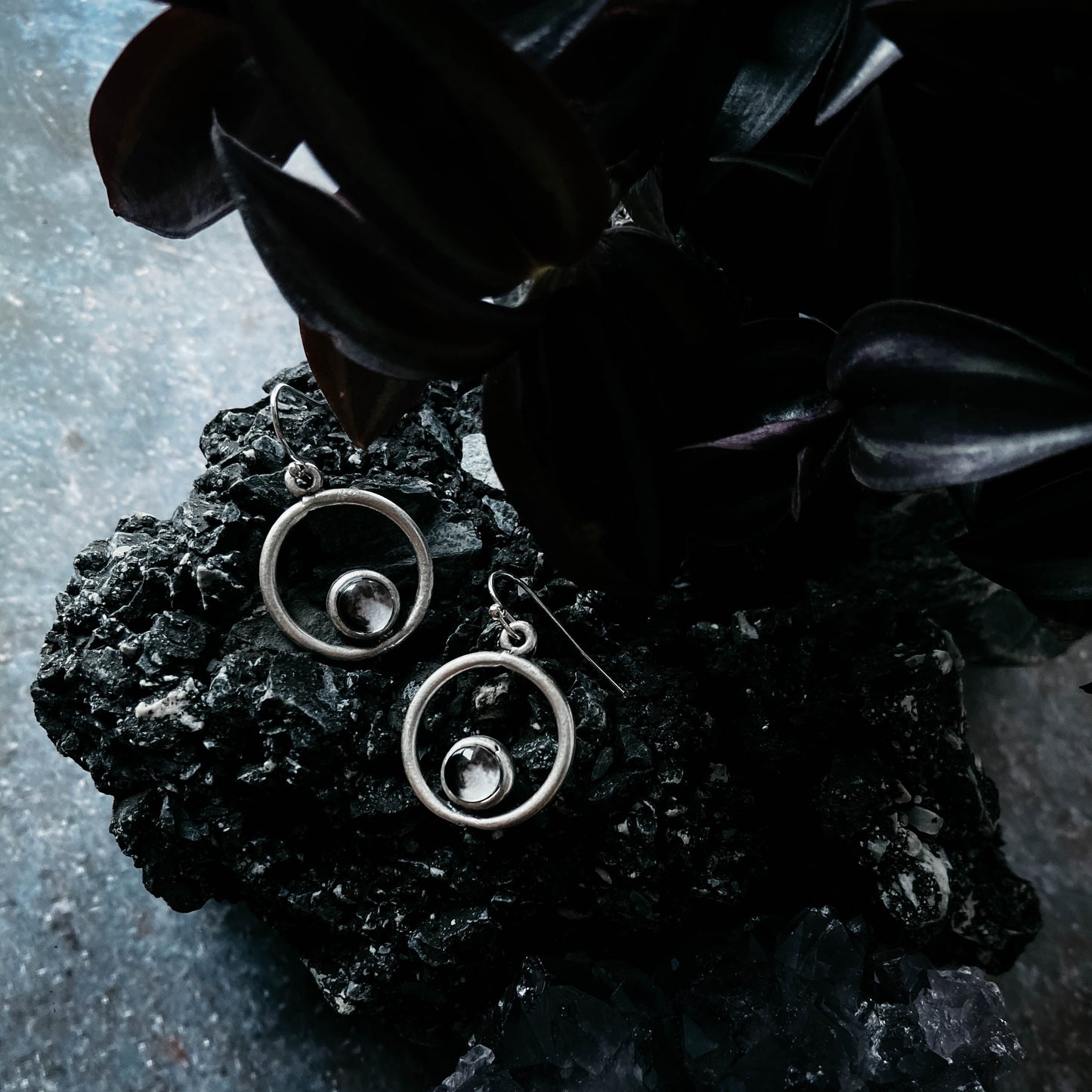 Silver circular earrings on a dark stone surface with leaves