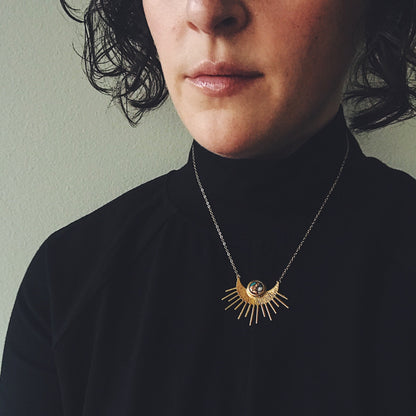 Person wearing a black turtleneck and a gold necklace with a sun design against a neutral background