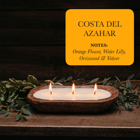 Three candles in a wooden holder with green leaves on a dark background, featuring 'Costa Del Azahar' scents.