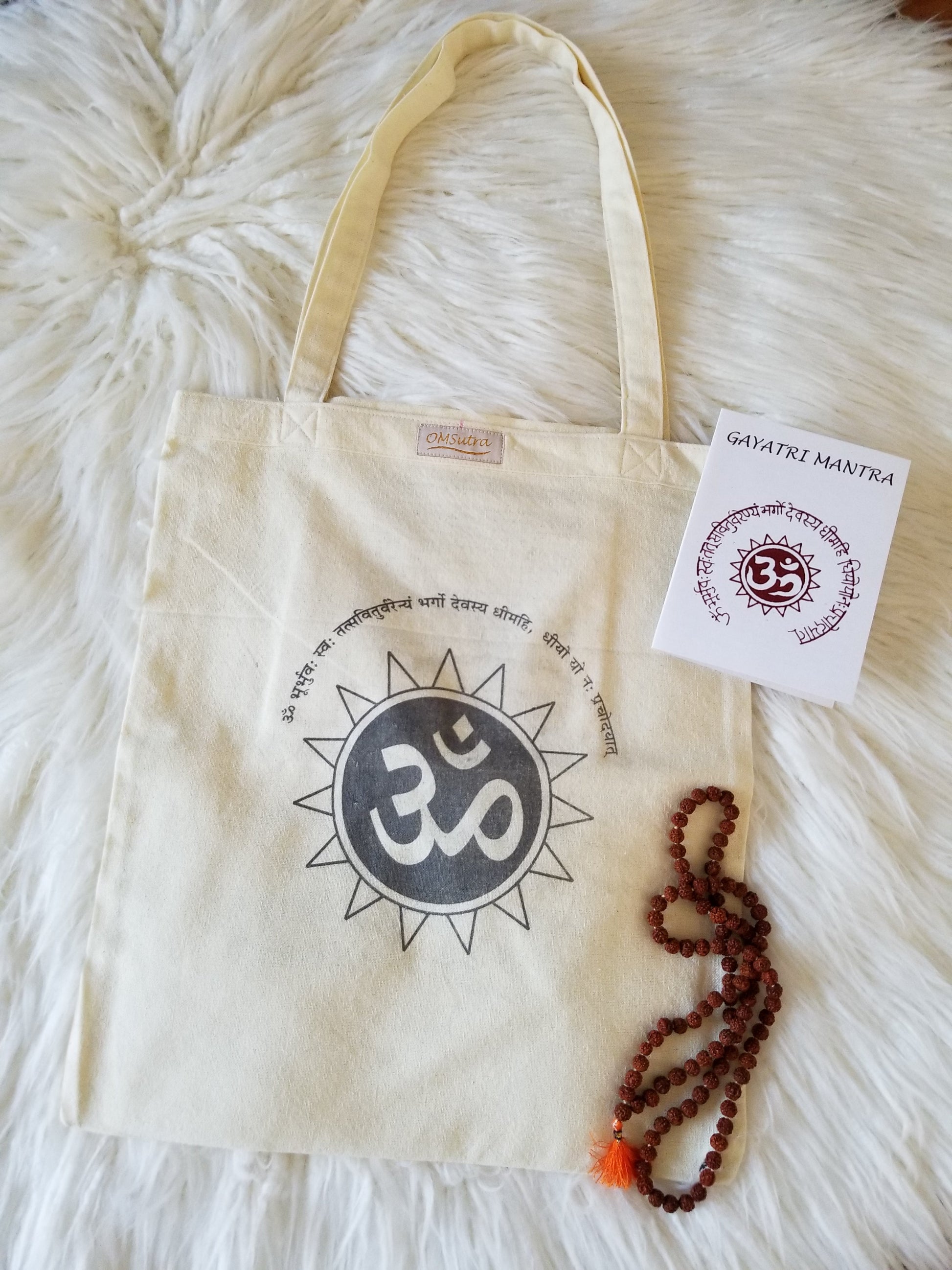 Beige tote bag with Om symbol, mala beads, and a card on a fluffy white surface