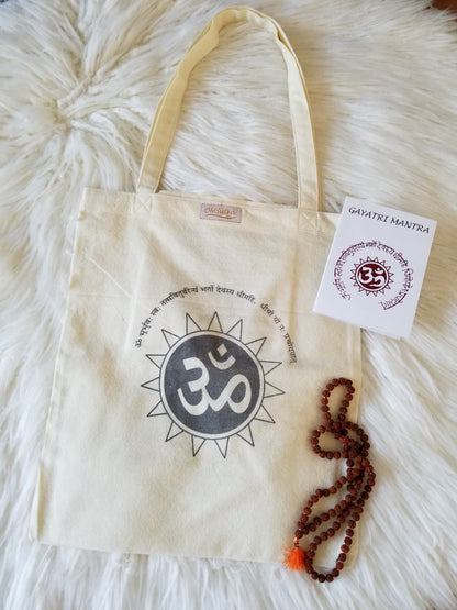 Beige tote bag with Om symbol, mala beads, and a card on a fluffy white surface