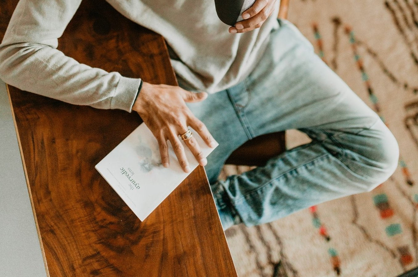 Person sitting at a wooden table with a book and a cup, wearing a light-colored sweater and jeans.
