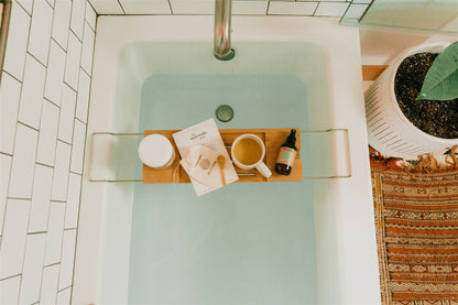 Bath tub with a tray holding a book, cup, and bottle, surrounded by a plant and towel.