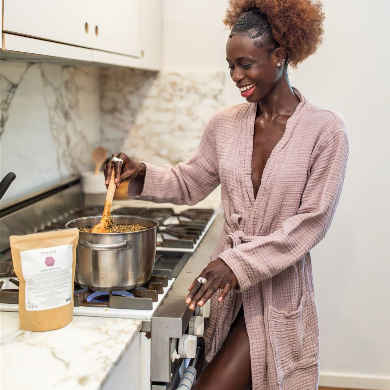 Woman in a pink robe cooking on a stove with a package of tea on the counter.
