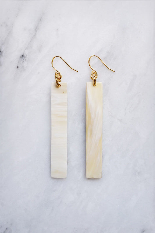 Pair of gold earrings with long, thin, beige stones on a marble background