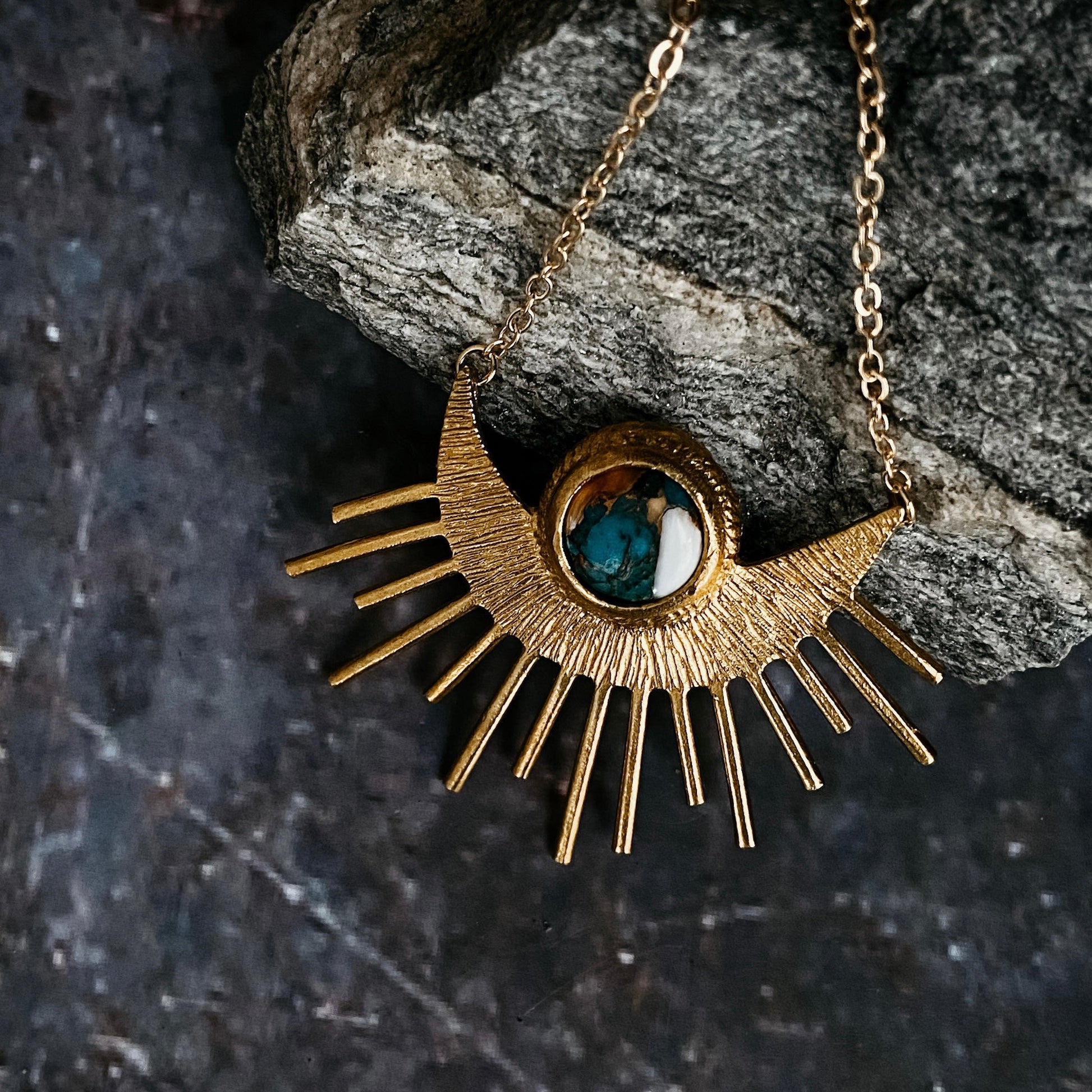 Gold necklace with a sun-shaped pendant featuring a blue gemstone on a dark stone background
