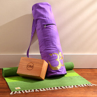 Purple yoga mat bag, wooden block, and green mat on a wooden floor.