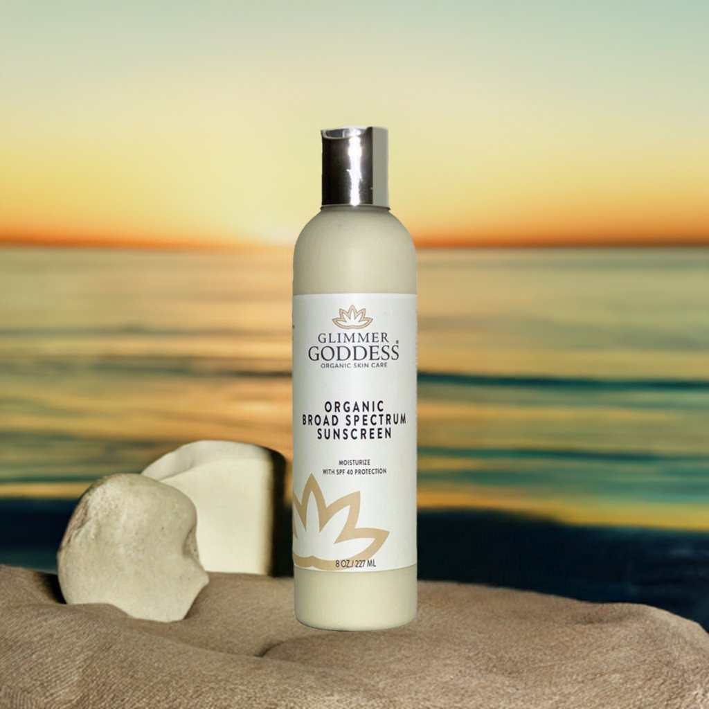 Bottle of Glimmer Goddess organic sunscreen on sand with a sunset background