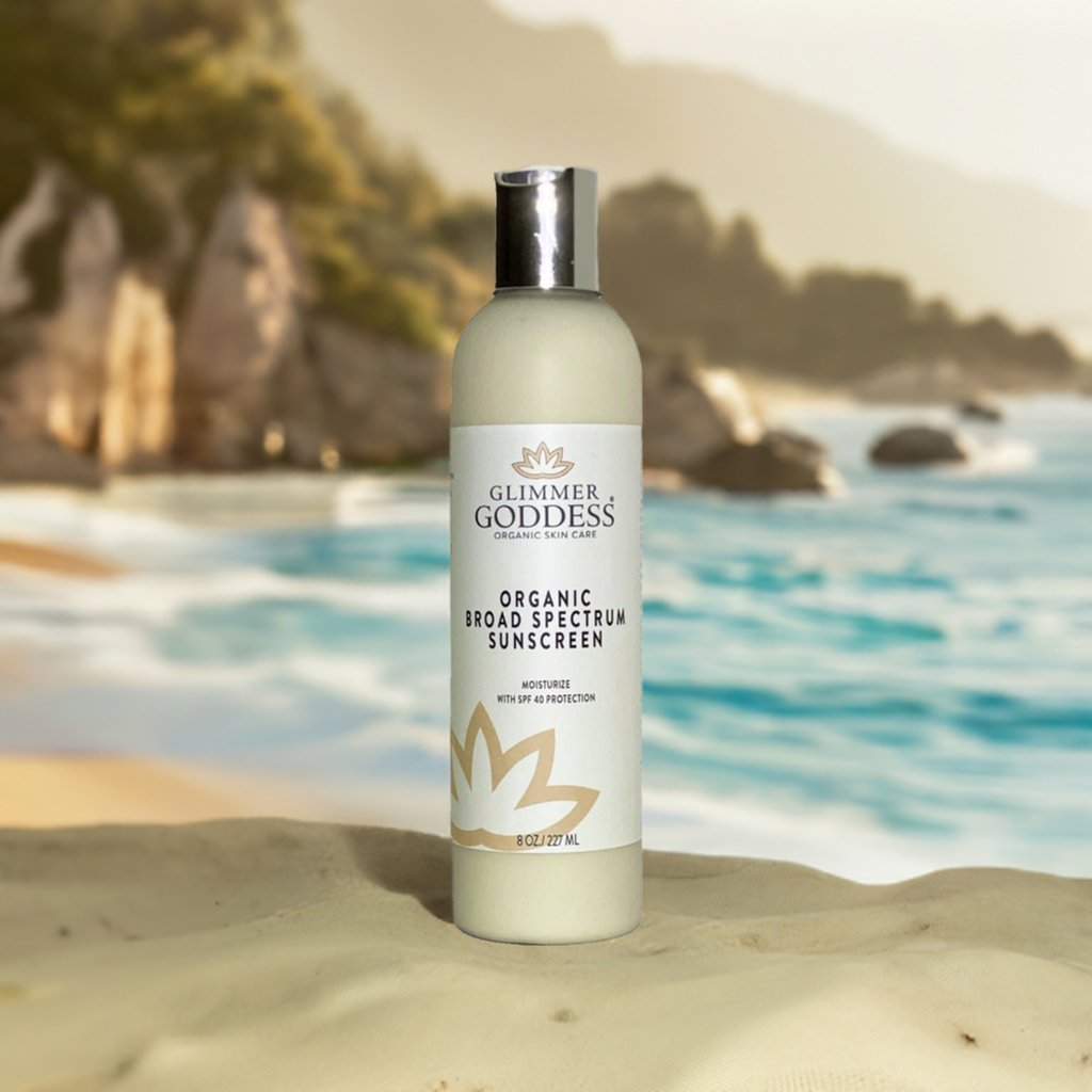Glimmer Goddess organic broad spectrum sunscreen bottle on a beach with ocean and rocks in the background