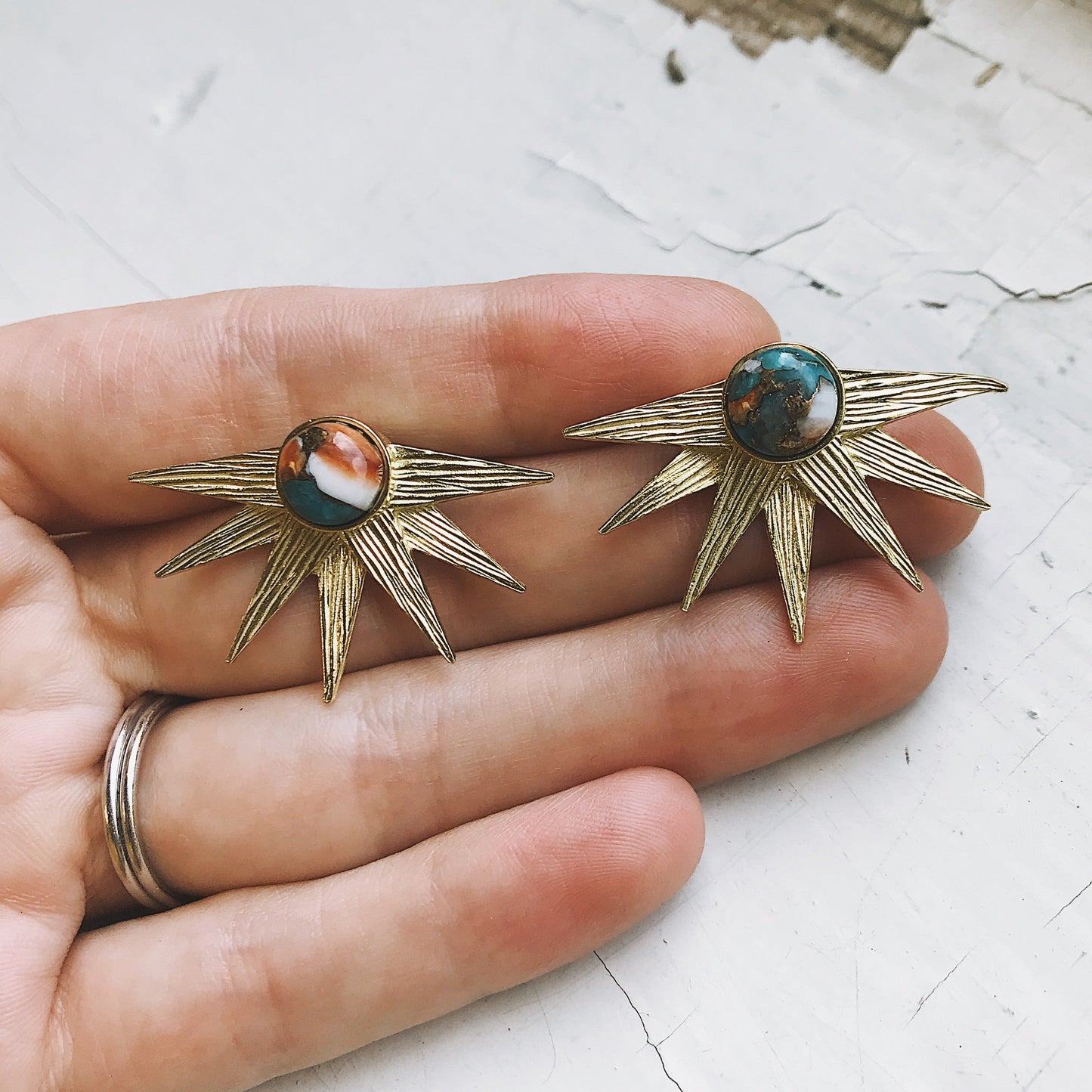 Gold sun-shaped earrings with gemstones held between fingers on a light background