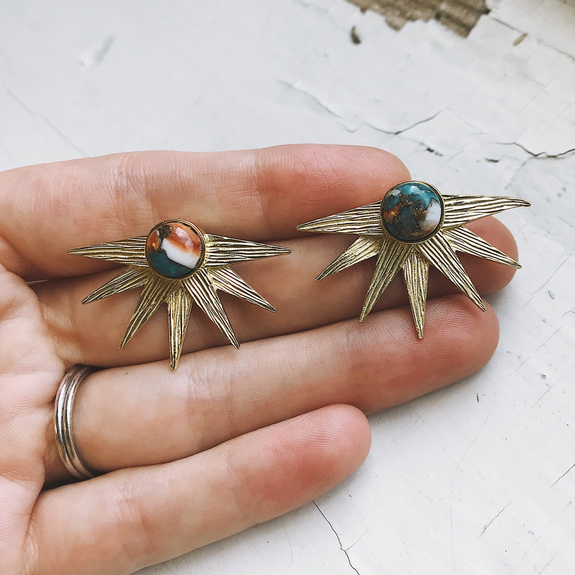 Gold sun-shaped earrings with gemstones held between fingers on a light background